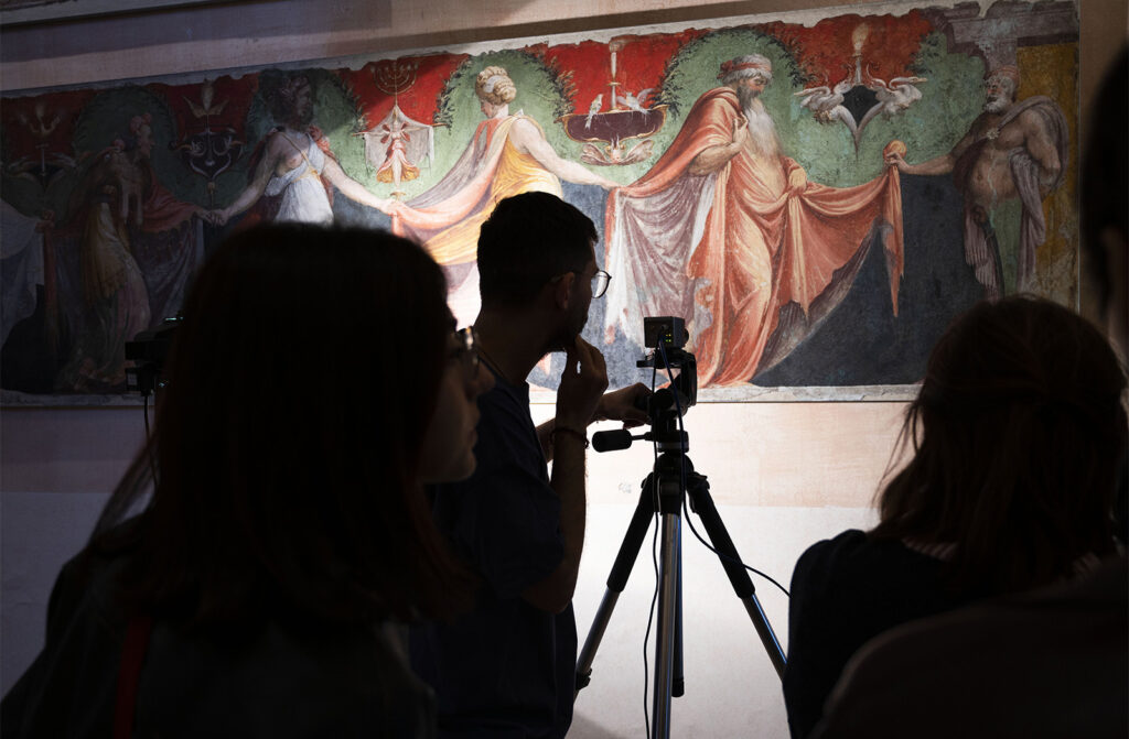 Gruppo di persone di fronte a un affresco colorato con figure umane; una persona utilizza una fotocamera montata su treppiede. Foto di Veronica Parrinello.